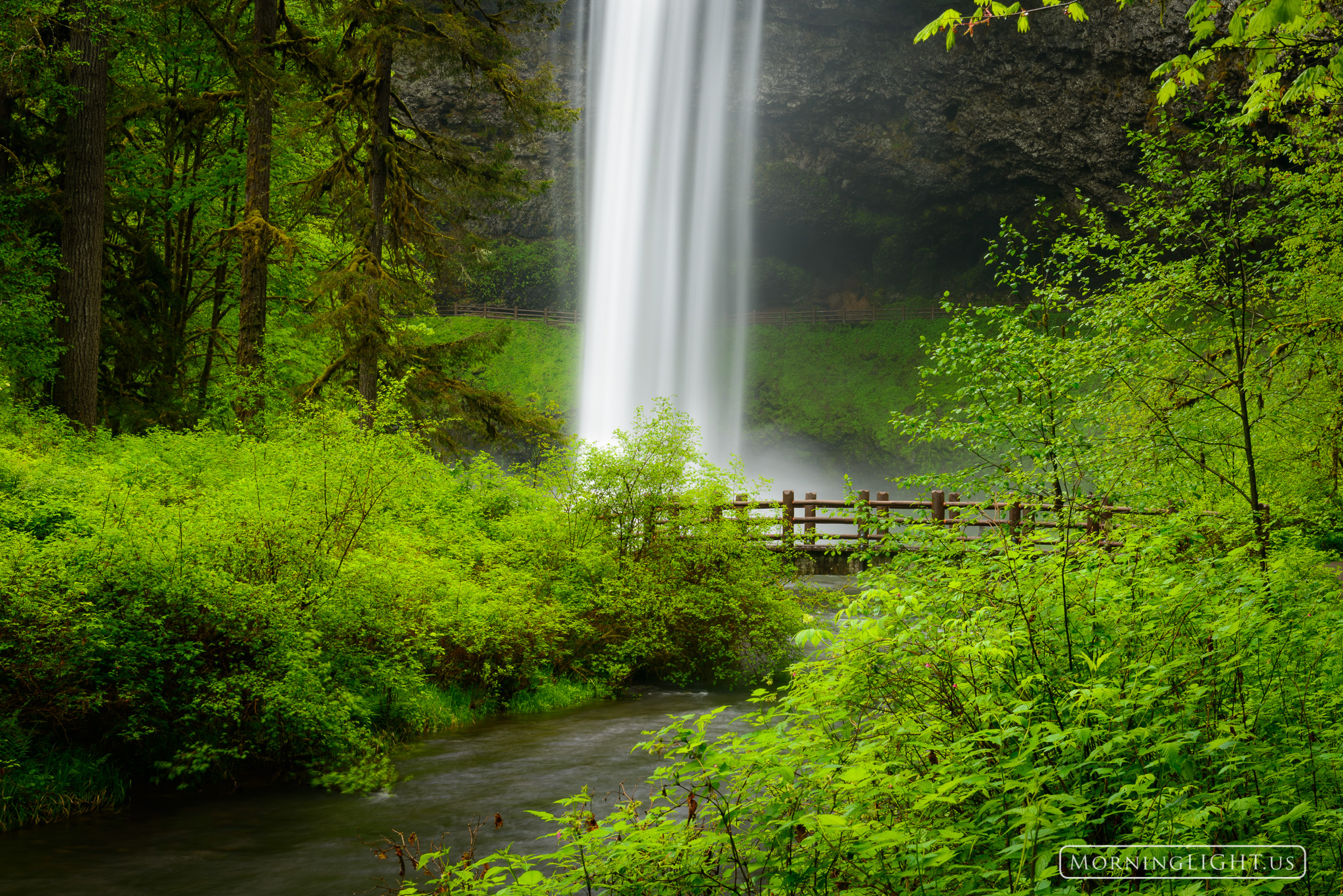 A waterfall comes out of the sky beside a small bridge in a lush Oregon forest.