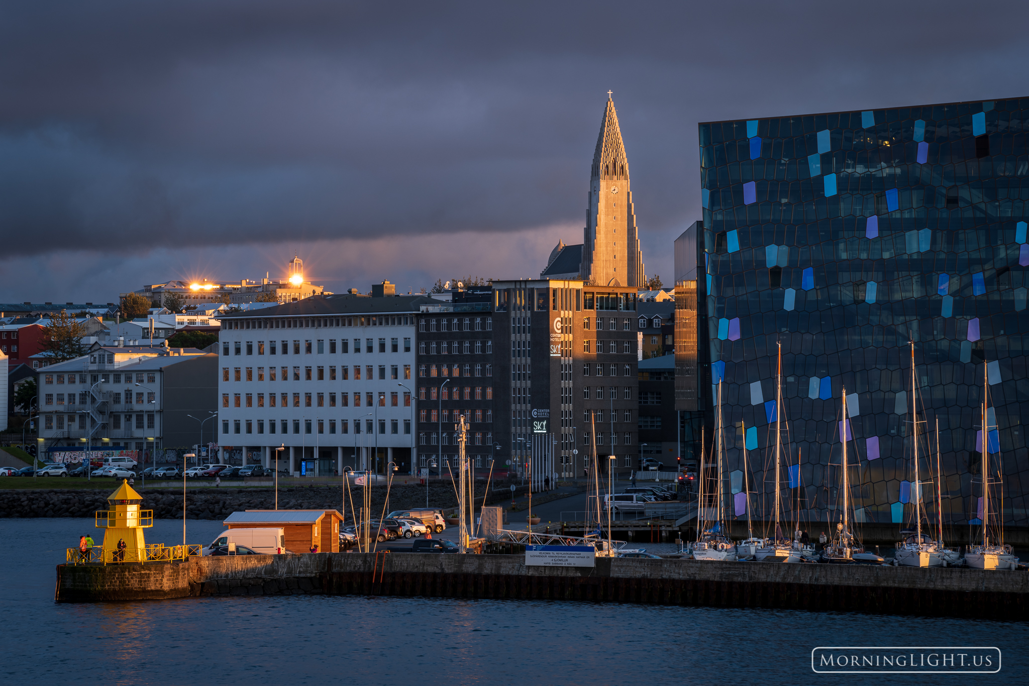 As the sun sets over the Atlantic Ocean, its warm light illuminates the spire of Hallgrimskirkja and the area surrounding Reykjav...